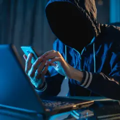 Osoba w kapturze korzysta z telefonu komórkowego przy laptopie w ciemnym pomieszczeniu.