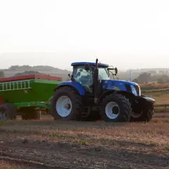 Niebieski traktor z przyczepą nawozową, gotowy do pracy na polu. Rolnik zastanawia się, jakie wapno na dofinansowanie będzie najlepsze.