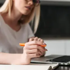 Kobieta z pomarańczowym długopisem w dłoni uczy się, jak napisać uzasadnienie wniosku o dofinansowanie, pracując przy laptopie.
