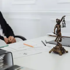 Prawnik przy biurku z dokumentami i figurką Temidy. Na stole leży laptop i stos dokumentów, w tym zapis bankowy.