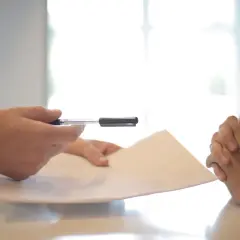 Dwie osoby przy stole, jedna z długopisem, druga z dłońmi złożonymi. Rozmowa o tym, jak sprawdzić najemcę w BIK.