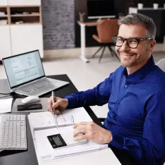Uśmiechnięty mężczyzna w okularach przy biurku z komputerem i dokumentami. Zastanawia się, ile można dostać dofinansowania na firmę.