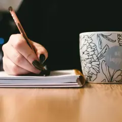 Dłoń pisze notatki, by przygotować się do napisania uzasadnienia wniosku o dofinansowanie komputera. Obok kubek z napisem "Be Happy".
