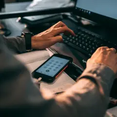 Ręce wpisujące kod na klawiaturze komputera, obok telefon z klawiaturą numeryczną. Może szukasz informacji, jak zmienić numer telefonu BLIK w mBank?