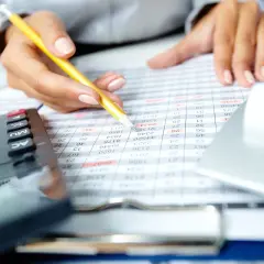 Ręka z ołówkiem analizuje tabelę finansową. Kalkulator i laptop w tle. Szukasz banku, który nie patrzy na zapytania w BIK?