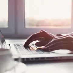 Dłonie piszą na laptopie, a obok stoi filiżanka kawy. Słońce wpada przez okno, tworząc ciepłą atmosferę. To idealny moment na przelew elixir.