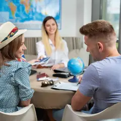 Para planować podróż, warto wiedzieć, co to turystyczny fundusz gwarancyjny. Para młodych ludzi z paszportami i globusem na stole omawia szczegóły wycieczki z pracownicą biura podróży.