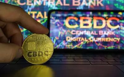 Złota moneta z napisem CBDC w dłoni, na tle ekranu z wykresem i napisem "Central Bank Digital Currency".