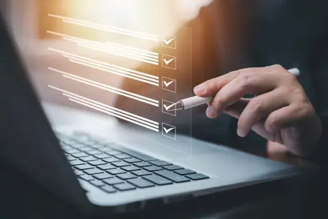 Ręka z długopisem zaznacza punkty na liście kontrolnej na ekranie laptopa. Proces zmiany regulaminu zakładowego funduszu świadczeń socjalnych.