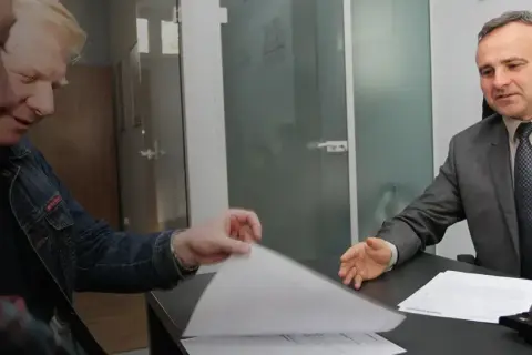 Mężczyzna w garniturze rozmawia z klientami przy biurku, przeglądając dokumenty.