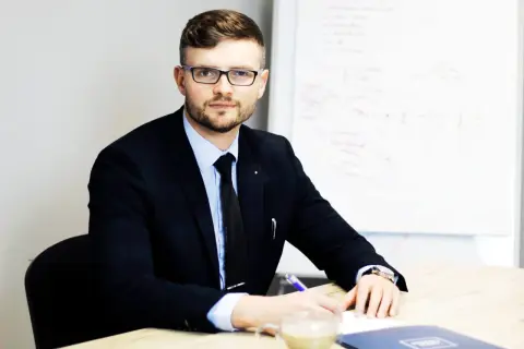 Mężczyzna w garniturze i okularach siedzi przy biurku, pisząc coś na kartce. Obok stoi filiżanka kawy.