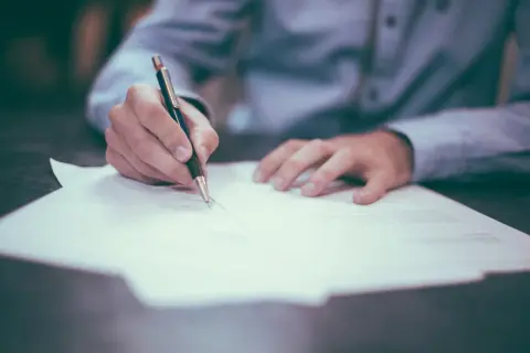 Ręka pisze długopisem na papierze. Proces sprawdzania BIK osoby zmarłej wymaga analizy dokumentów.