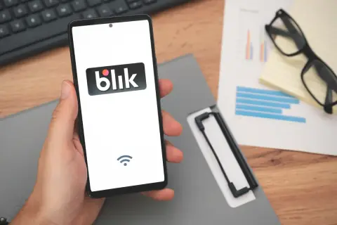 Dłoń trzyma smartfon z logo Blik, sugerując szybki zarobek. Na biurku leżą dokumenty i okulary.