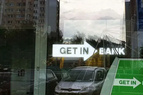 Czy lokaty w Getin Banku są bezpieczne? Znak "GET IN BANK" odbija się w szybie, obok samochodów i budynków.