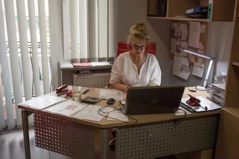 Kobieta w okularach pracuje przy biurku z laptopem, dokumentami i kalkulatorem.