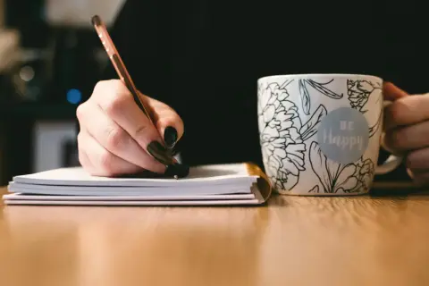 Dłoń pisze notatki, by przygotować się do napisania uzasadnienia wniosku o dofinansowanie komputera. Obok kubek z napisem "Be Happy".