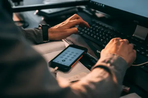 Ręce wpisujące kod na klawiaturze komputera, obok telefon z klawiaturą numeryczną. Może szukasz informacji, jak zmienić numer telefonu BLIK w mBank?