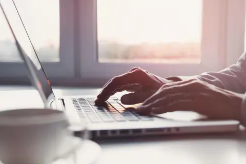 Dłonie piszą na laptopie, a obok stoi filiżanka kawy. Słońce wpada przez okno, tworząc ciepłą atmosferę. To idealny moment na przelew elixir.