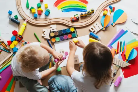Dzieci tworzą kolorowe prace, jakby planowały, jak zdobyć dofinansowanie do przedszkola. Widać klocki, farby, kredki i papier.