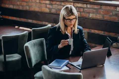 Kobieta w okularach pracuje na laptopie, popijając kawę. Na stole leży notatnik, być może planuje swój **konto Aurum**.