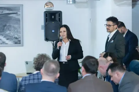 Kobieta w czarnym żakiecie przemawia do publiczności, trzymając mikrofon.