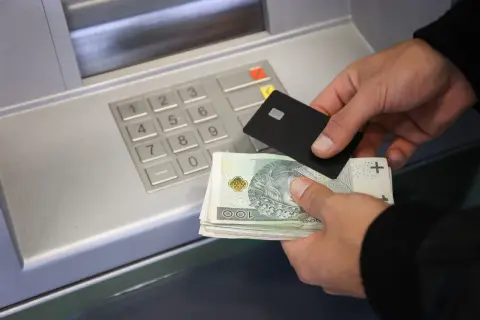 Ręce trzymające banknoty i kartę zbliżają się do bankomatu, pokazując, jak wpłacić pieniądze na konto.