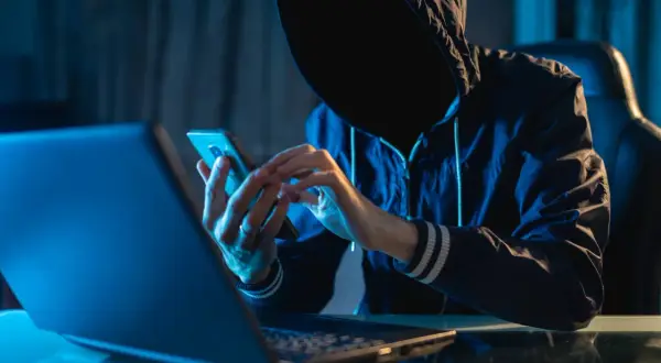 Osoba w kapturze korzysta z telefonu komórkowego przy laptopie w ciemnym pomieszczeniu.