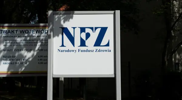 Tablica z logo NFZ i napisem Narodowy Fundusz Zdrowia. Obok tablica z informacjami o inwestycji w szpital.
