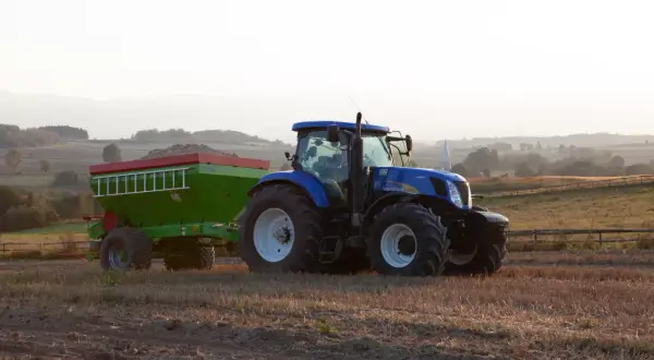 Niebieski traktor z przyczepą nawozową, gotowy do pracy na polu. Rolnik zastanawia się, jakie wapno na dofinansowanie będzie najlepsze.