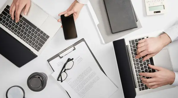Dwie osoby pracują przy biurku, używając laptopów i telefonu. Na stole leżą dokumenty, okulary, lupa i kubek. Sprawdzają, ile widnieje się w BIK.
