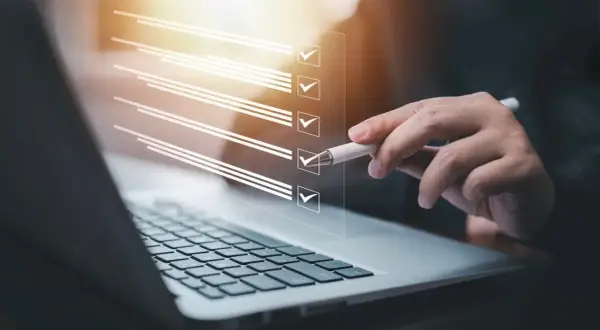 Ręka z długopisem zaznacza punkty na liście kontrolnej na ekranie laptopa. Proces zmiany regulaminu zakładowego funduszu świadczeń socjalnych.