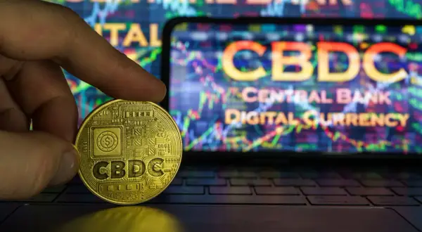 Złota moneta z napisem CBDC w dłoni, na tle ekranu z wykresem i napisem "Central Bank Digital Currency".