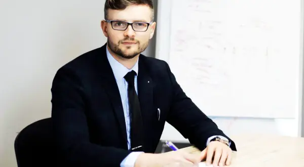 Mężczyzna w garniturze i okularach siedzi przy biurku, pisząc coś na kartce. Obok stoi filiżanka kawy.