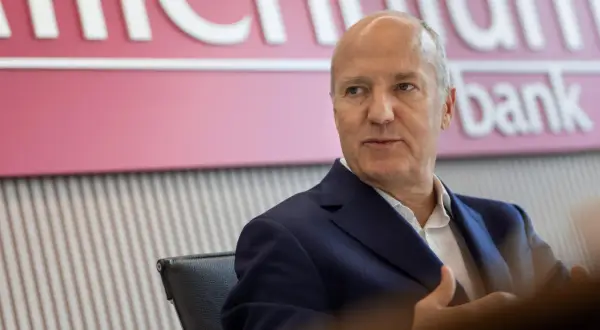 Mężczyzna w garniturze siedzi przed logo Banku Millennium. Kto jest właścicielem banku Millennium?