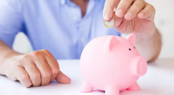 Mężczyzna wrzuca monetę do różowej świnki-skarbonki, zastanawiając się, czy lepsze będą lokaty czy konto oszczędnościowe.