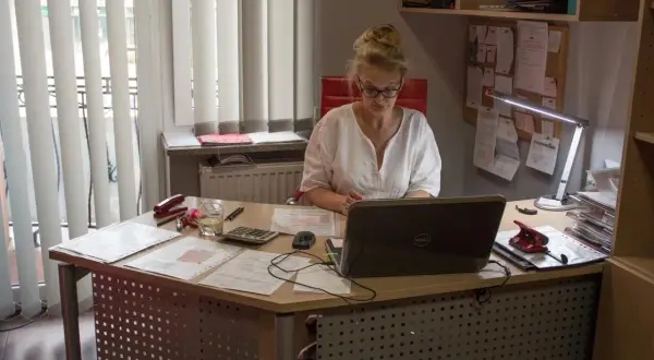 Kobieta w okularach pracuje przy biurku z laptopem, dokumentami i kalkulatorem.