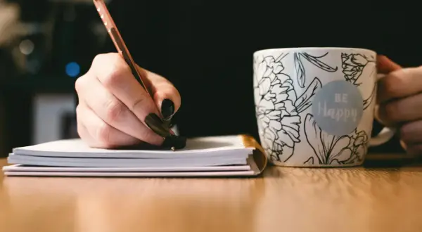 Dłoń pisze notatki, by przygotować się do napisania uzasadnienia wniosku o dofinansowanie komputera. Obok kubek z napisem "Be Happy".