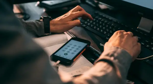Ręce wpisujące kod na klawiaturze komputera, obok telefon z klawiaturą numeryczną. Może szukasz informacji, jak zmienić numer telefonu BLIK w mBank?