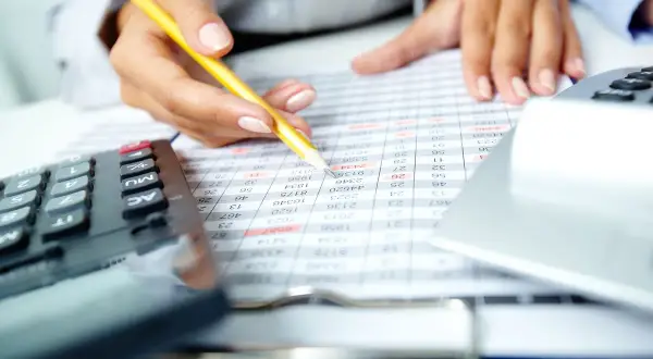 Ręka z ołówkiem analizuje tabelę finansową. Kalkulator i laptop w tle. Szukasz banku, który nie patrzy na zapytania w BIK?