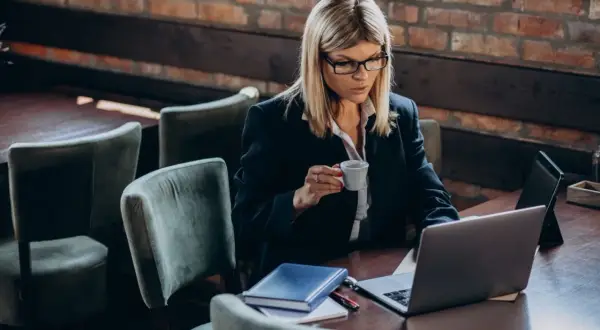 Kobieta w okularach pracuje na laptopie, popijając kawę. Na stole leży notatnik, być może planuje swój **konto Aurum**.