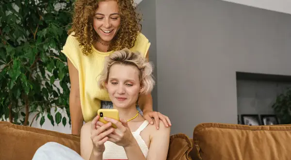 Dwie kobiety śmieją się, przeglądając telefon. Jedna z nich kładzie dłoń na ramieniu drugiej, sugerując wspólne konto dla par.