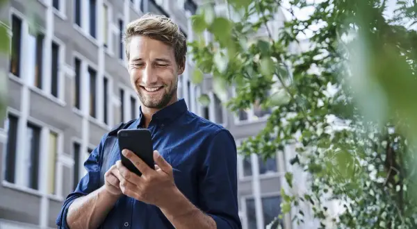 Uśmiechnięty mężczyzna sprawdza telefon, zastanawiając się, czy lokata mobilna jest bezpieczna. W tle budynek i zieleń.