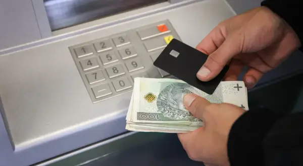 Ręce trzymające banknoty i kartę zbliżają się do bankomatu, pokazując, jak wpłacić pieniądze na konto.