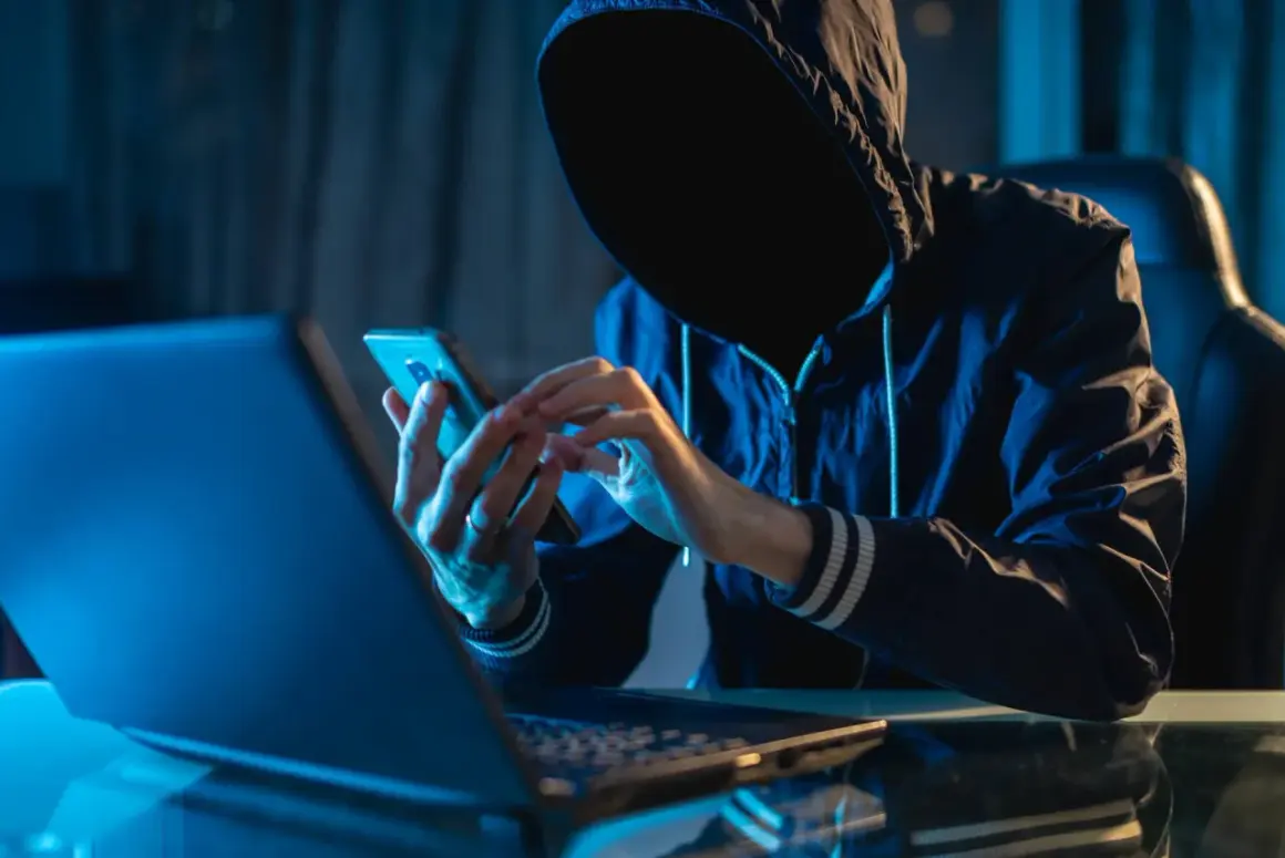 Osoba w kapturze korzysta z telefonu komórkowego przy laptopie w ciemnym pomieszczeniu.