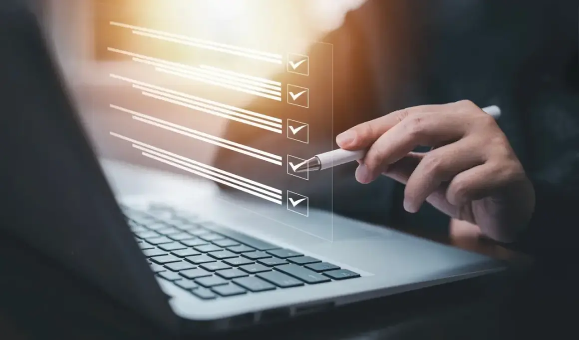 Ręka z długopisem zaznacza punkty na liście kontrolnej na ekranie laptopa. Proces zmiany regulaminu zakładowego funduszu świadczeń socjalnych.