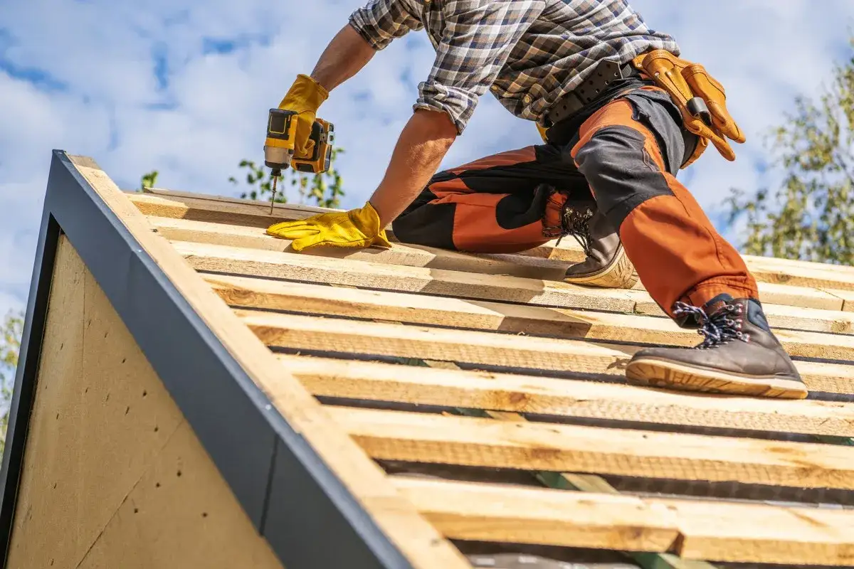 Budowa dachu to dobry moment, by dowiedzieć się, jak uzyskać dofinansowanie do wymiany dachu. Pracownik w żółtych rękawicach przykręca deski.