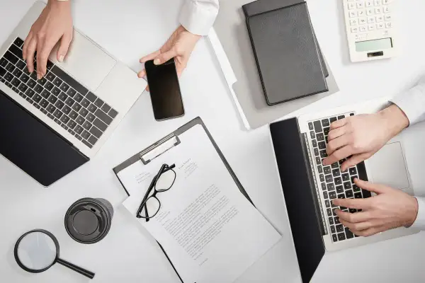 Dwie osoby pracują przy biurku, używając laptopów i telefonu. Na stole leżą dokumenty, okulary, lupa i kubek. Sprawdzają, ile widnieje się w BIK.