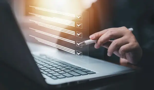 Ręka z długopisem zaznacza punkty na liście kontrolnej na ekranie laptopa. Proces zmiany regulaminu zakładowego funduszu świadczeń socjalnych.