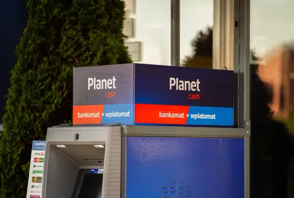 Bankomat Planet Cash z funkcją wpłatomatu, otoczony zielenią.