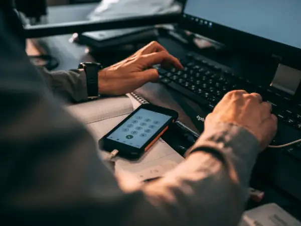 Ręce wpisujące kod na klawiaturze komputera, obok telefon z klawiaturą numeryczną. Może szukasz informacji, jak zmienić numer telefonu BLIK w mBank?